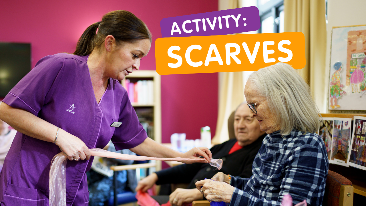 A white female care staff member holds a scarf with an elderly white woman who lives in the care home